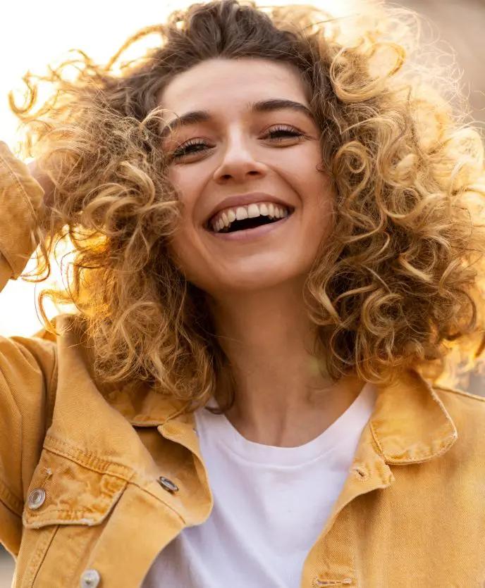 Woman with curly hair laughing outside wearing a yellow jacket.