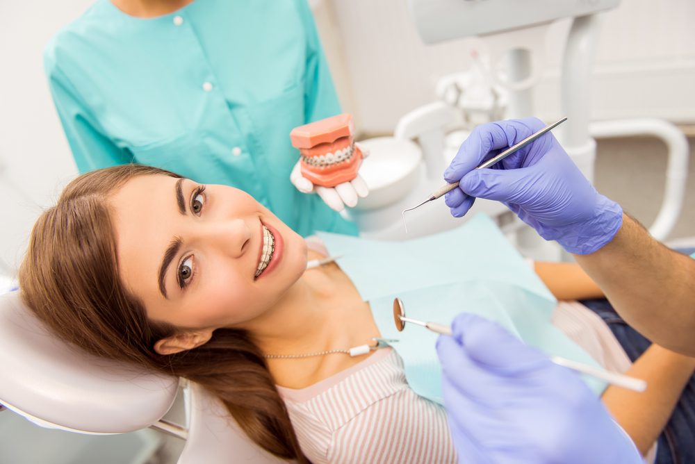 Attractive patient, with braces, treatment in dental, assistant to holding the model teeth, smiling, looking at camera.
